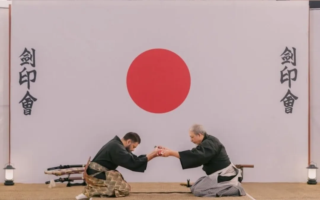 Two swordsmen in hakama kneeling to exchange a sword before a red rising-sun backdrop with Keninkai calligraphy